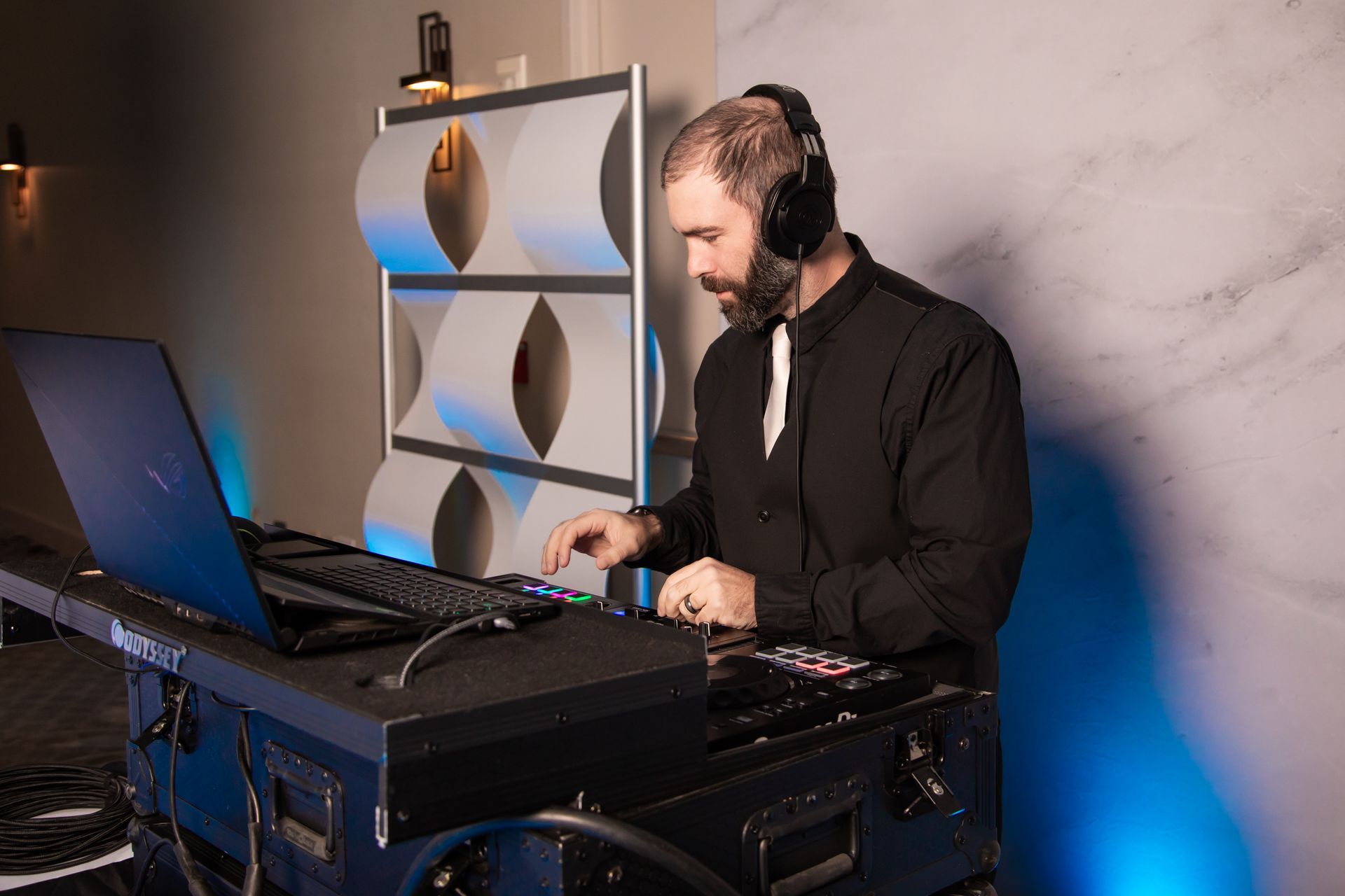 A man wearing headphones operates music equipment, including a laptop and keyboard, in front of a decorative wall.