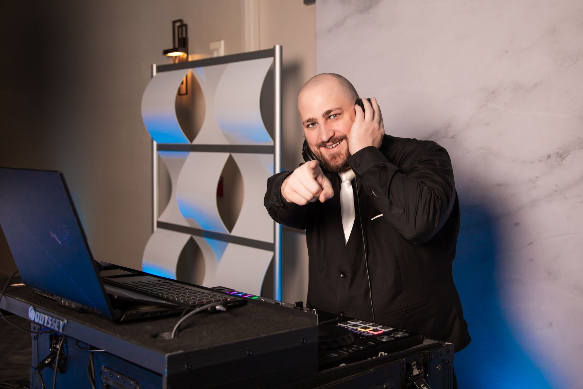DJ smiling, pointing, and wearing headphones next to laptop and music equipment. Blue and grey backdrop.