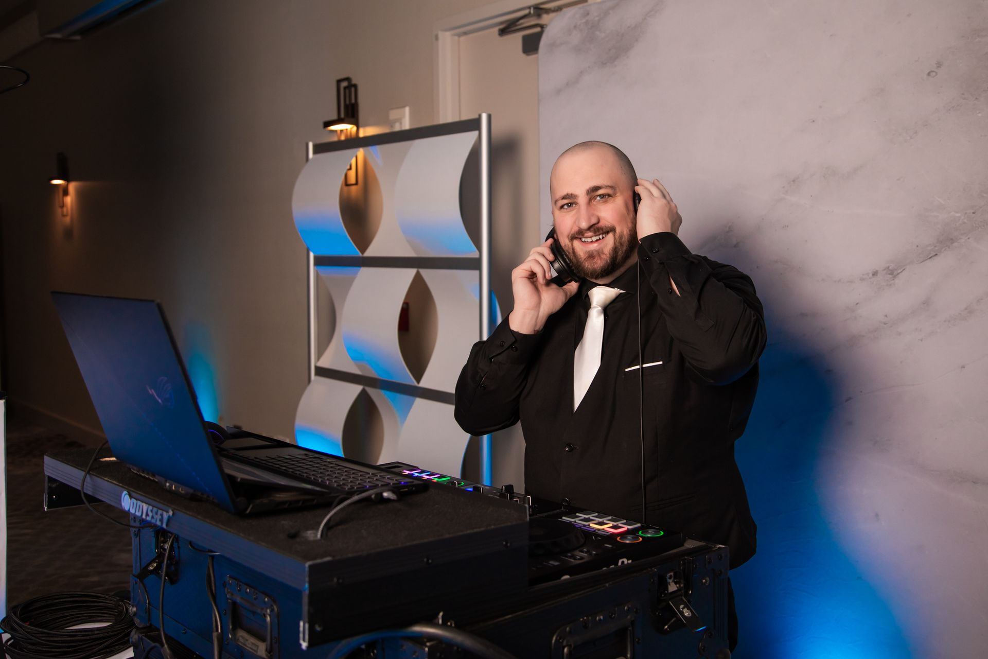 DJ with headphones smiles, standing near his equipment in front of a decorative wall.