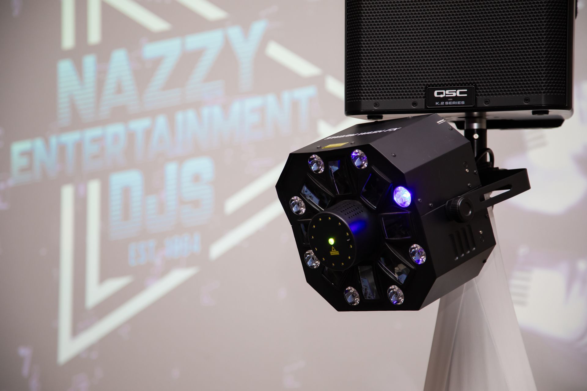 Black DJ lighting fixture with blue and green lights, next to a black speaker. The logo