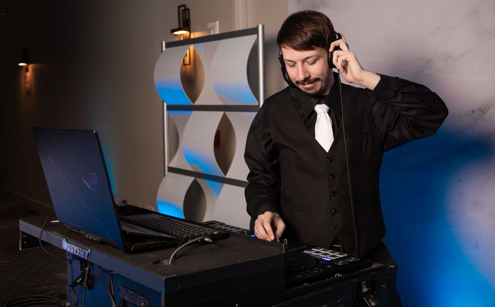 DJ wearing headphones, adjusting equipment behind a laptop, with a patterned backdrop.