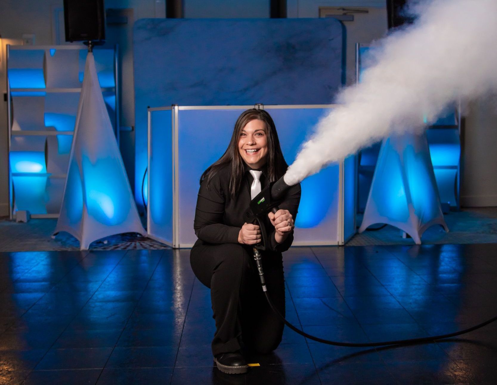 Woman kneeling, holding a fog machine that emits a cloud of smoke in a blue-lit setting.