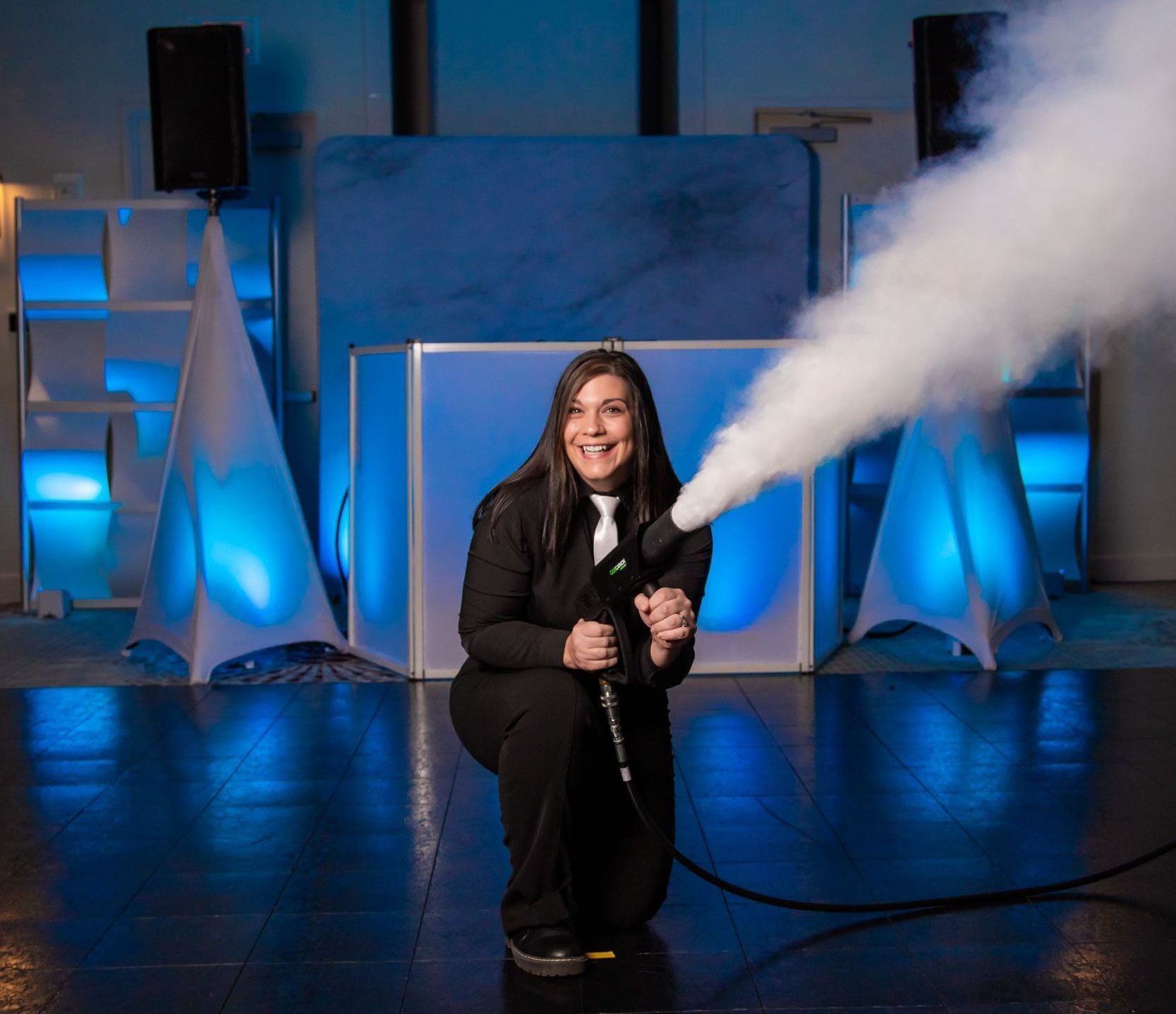 Woman kneeling, operating a smoke machine, smiling, in a blue-lit event space.