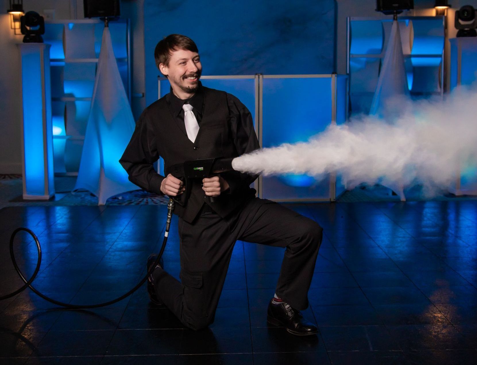 Man in formal wear kneels, aiming a smoke machine. Smoke billows out. Blue lights and stage backdrop.