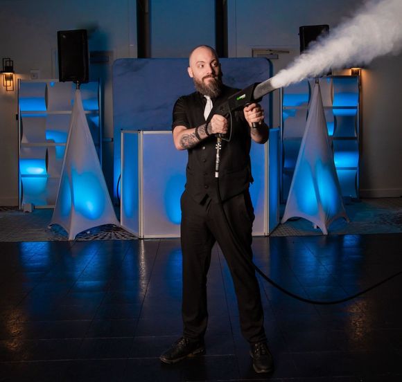 Man in black outfit holds fog machine, aiming it in room with blue stage lighting.