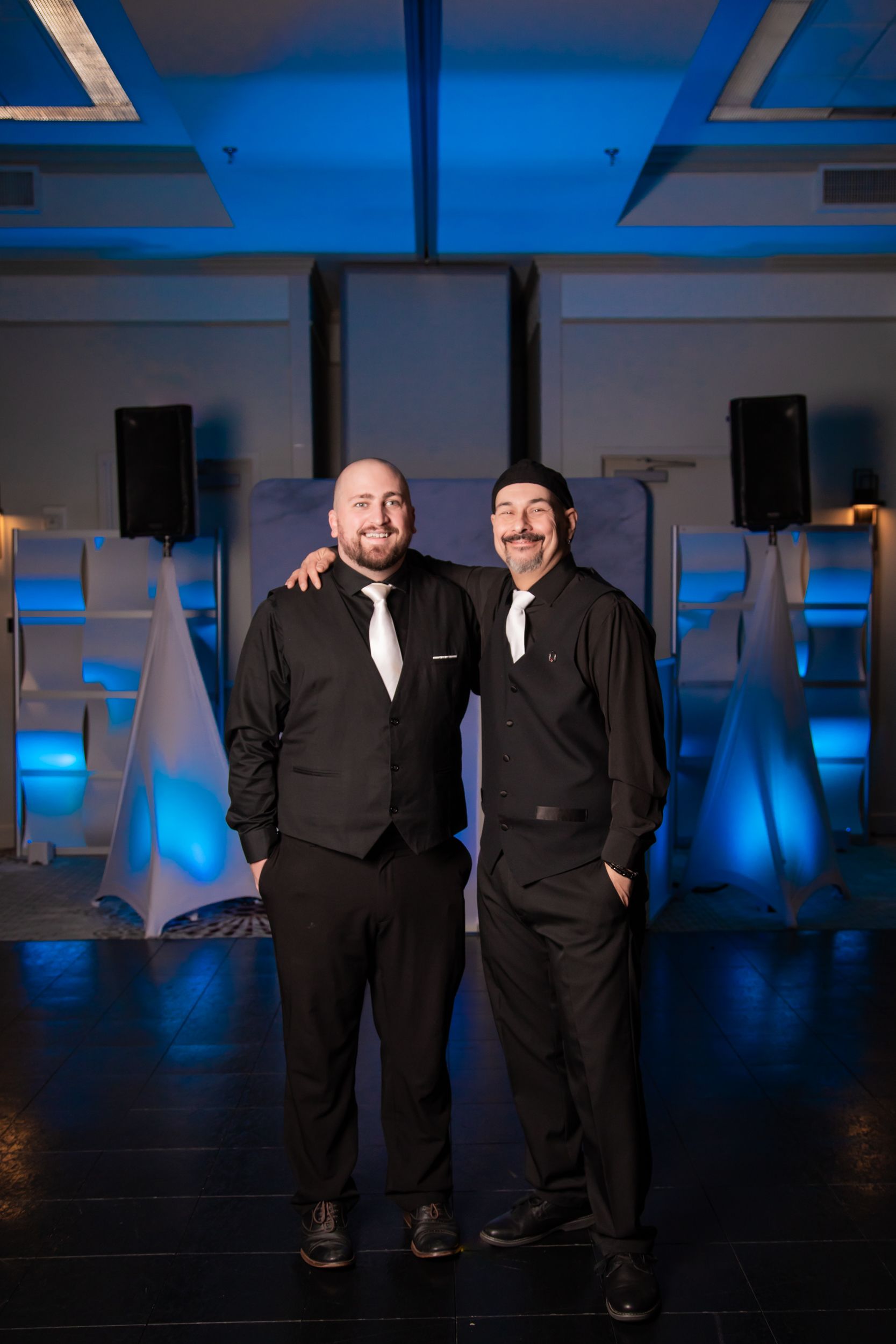 Two men in black vests and dress shirts pose with arms around each other in a blue-lit event space.
