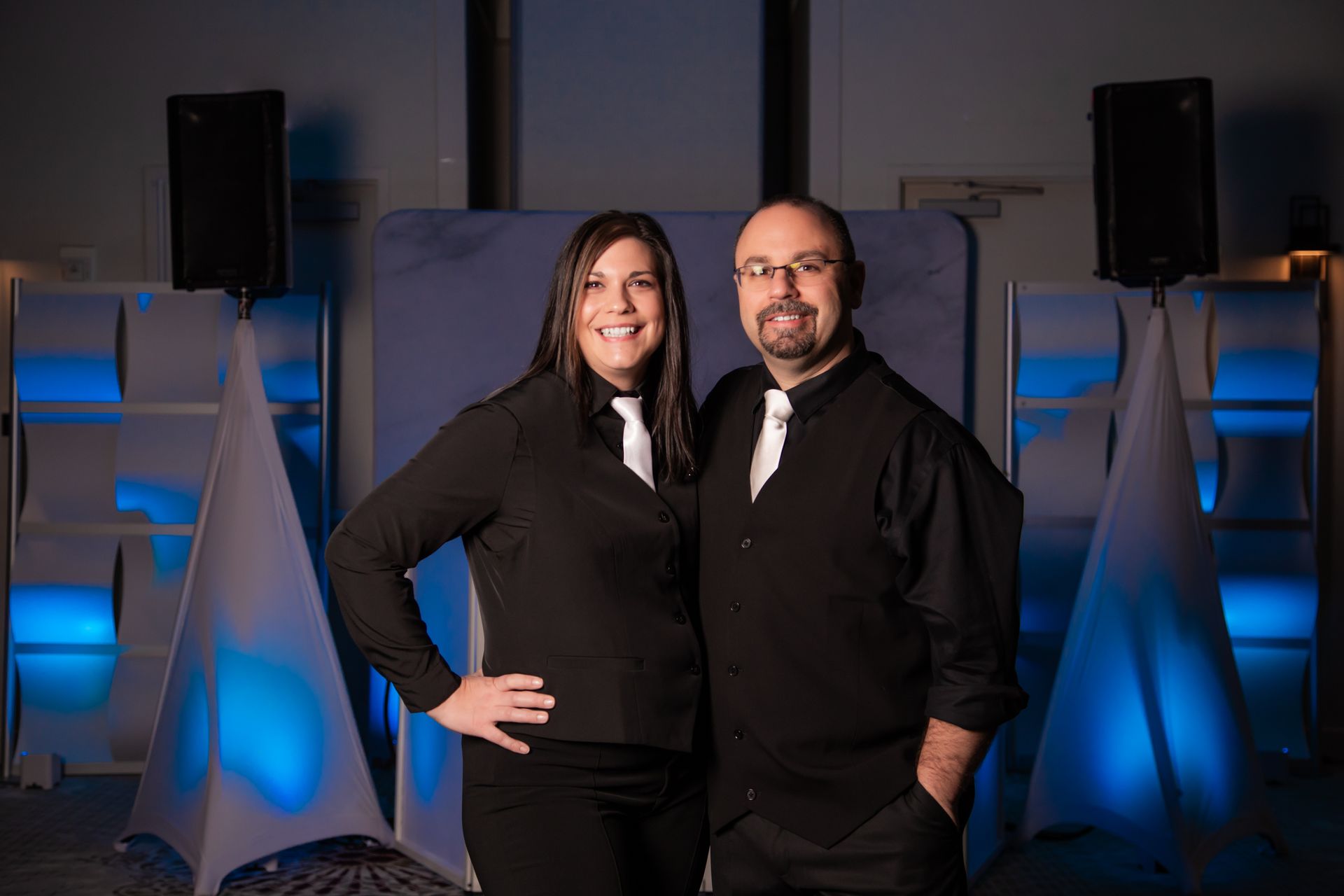 Two people in black attire pose in front of a blue-lit stage setup.