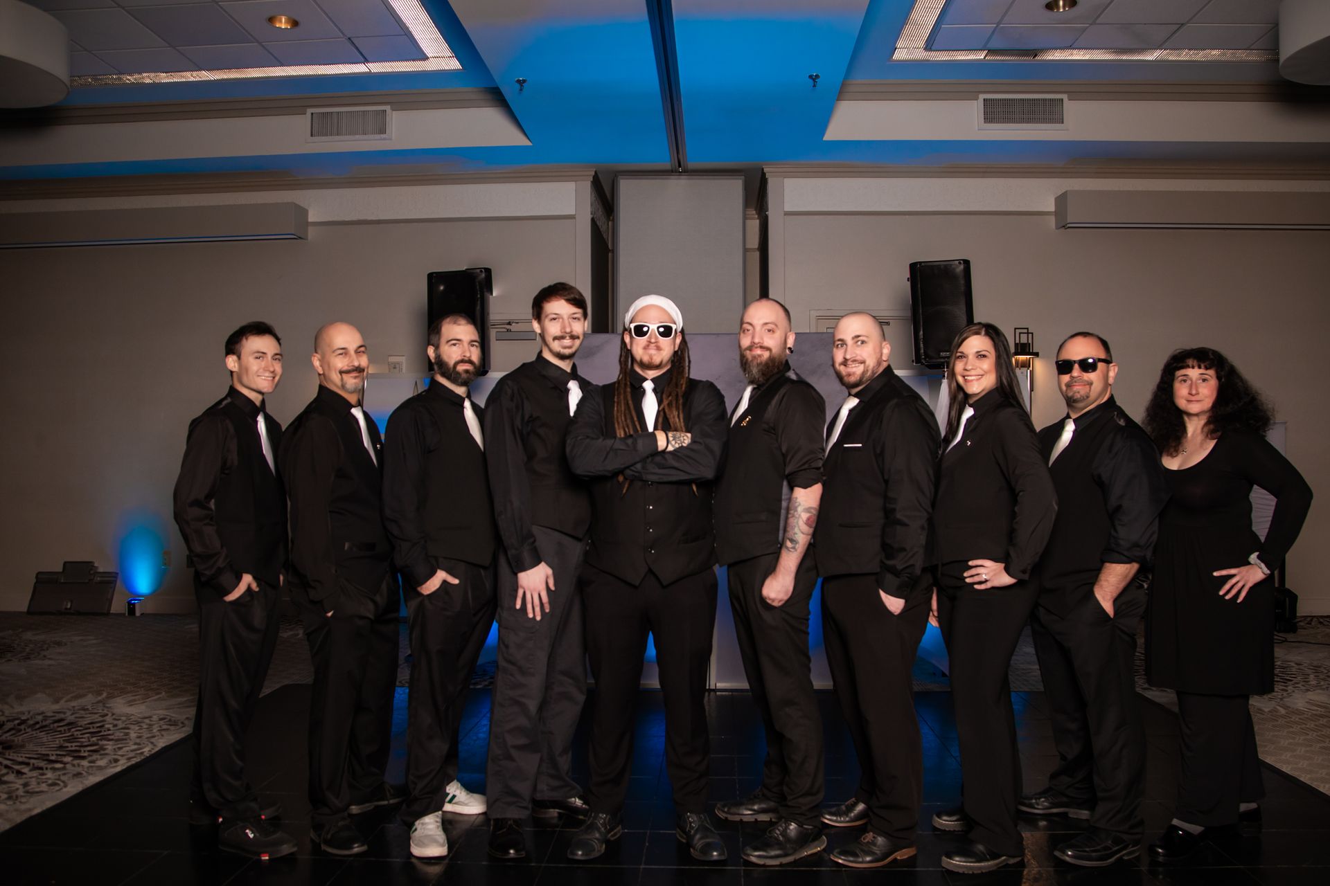Ten musicians in black suits pose indoors under blue lights.