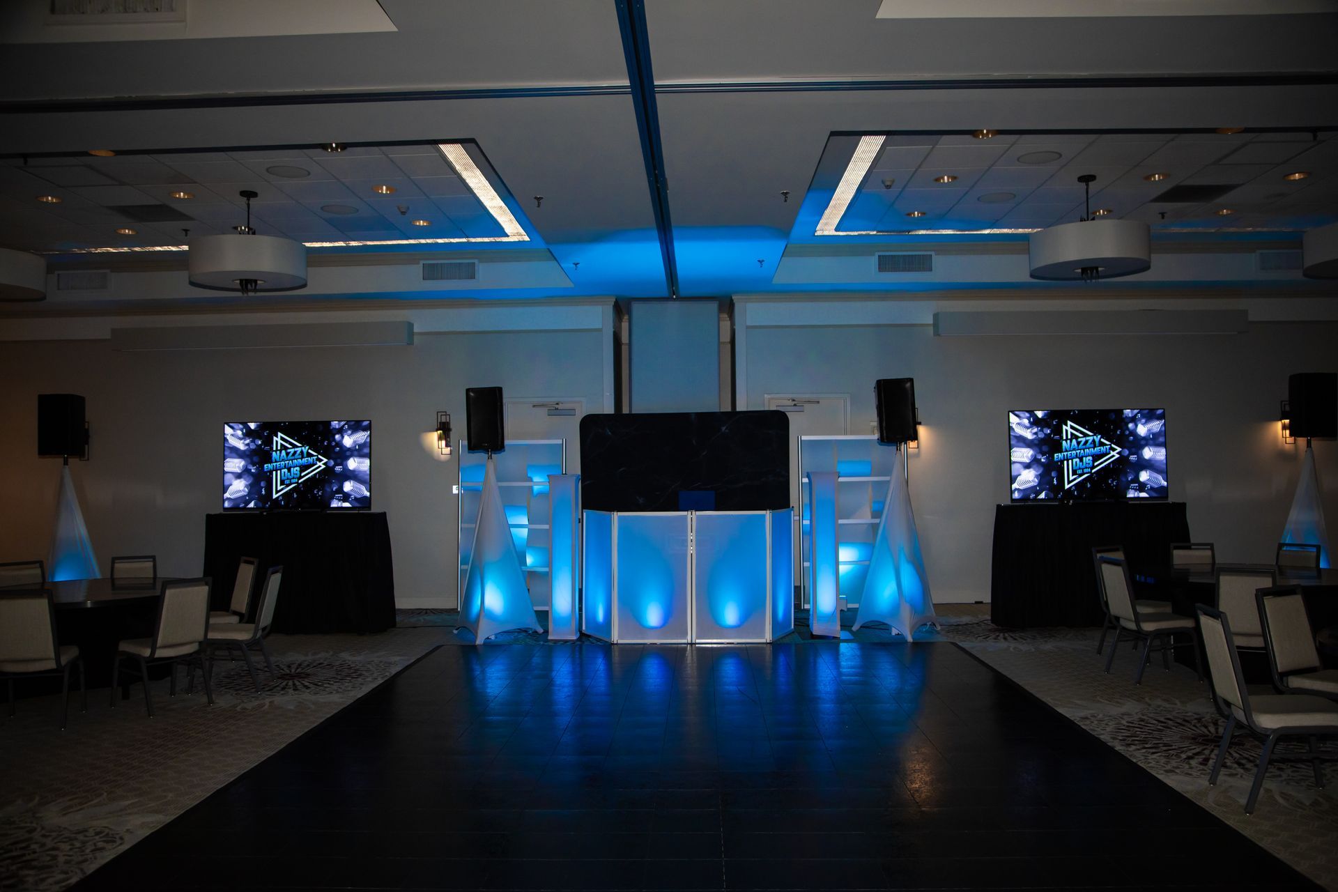 Blue-lit DJ setup in a ballroom. Two screens flank the DJ booth. Black dance floor, tables, and chairs.