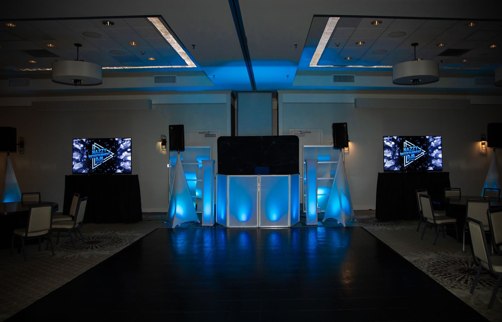 DJ setup with screens, speakers, and blue lighting in a large room with a dance floor.