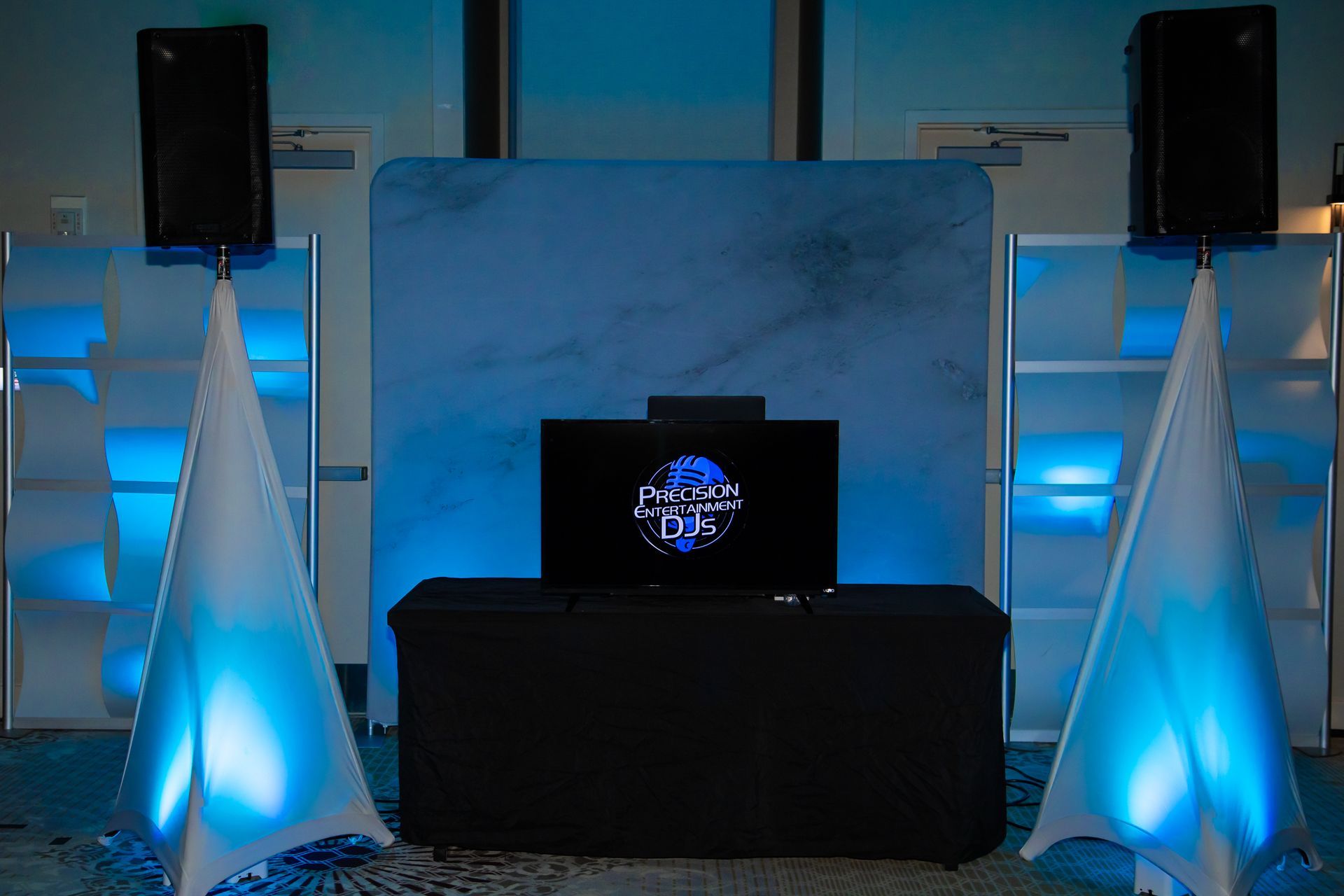 DJ setup with speakers, illuminated backdrop, and table with screen, bathed in blue light.