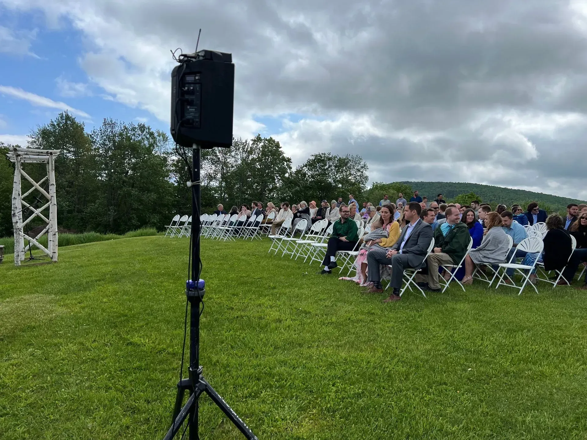 A sound system set up for an outdoor wedding. Guests seated on white chairs on a grassy hill.