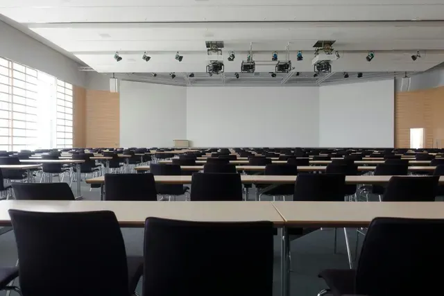 Rows of empty desks and chairs face a stage with a large white screen in a conference room.