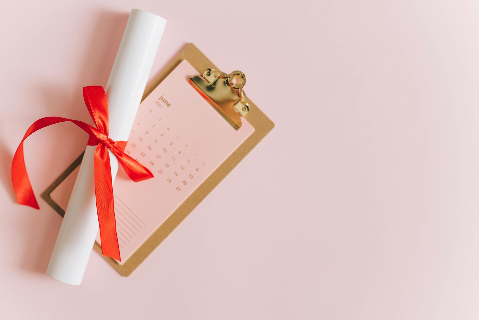 Diploma tied with red ribbon next to a clipboard with a pink calendar on a pink background.