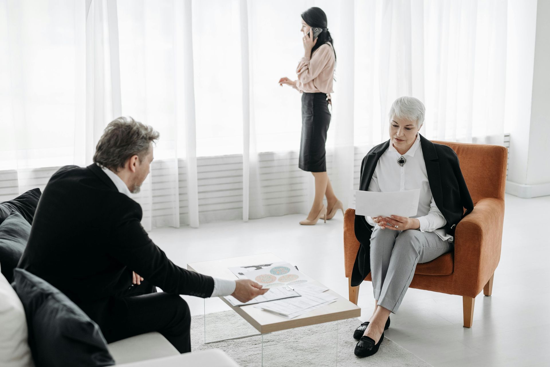 Three people in a bright office: a man gestures at documents, a woman reviews paperwork, and a third talks on the phone.