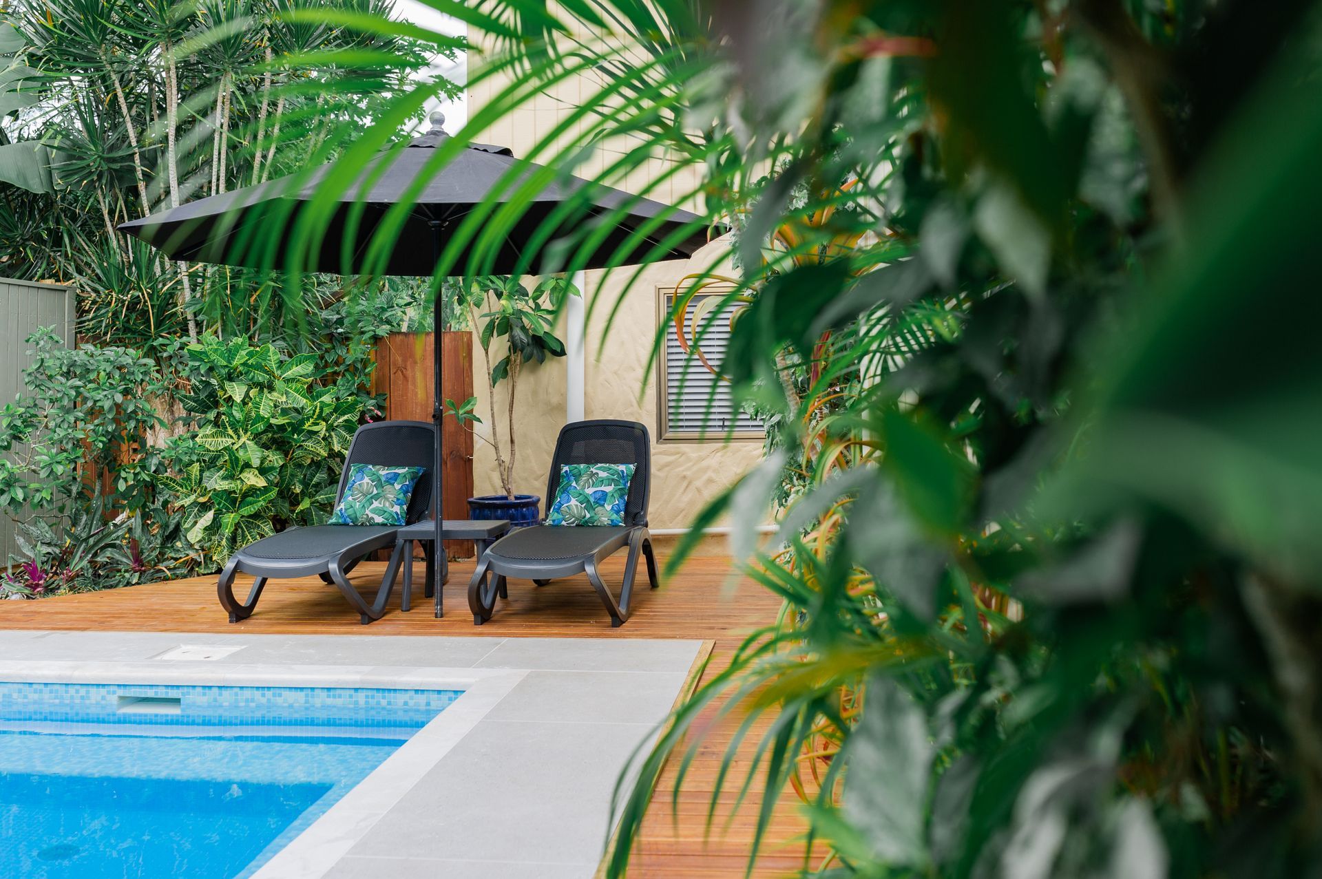 Two lounge chairs with patterned pillows under a black umbrella on a wooden deck by a swimming pool, surrounded by plants.