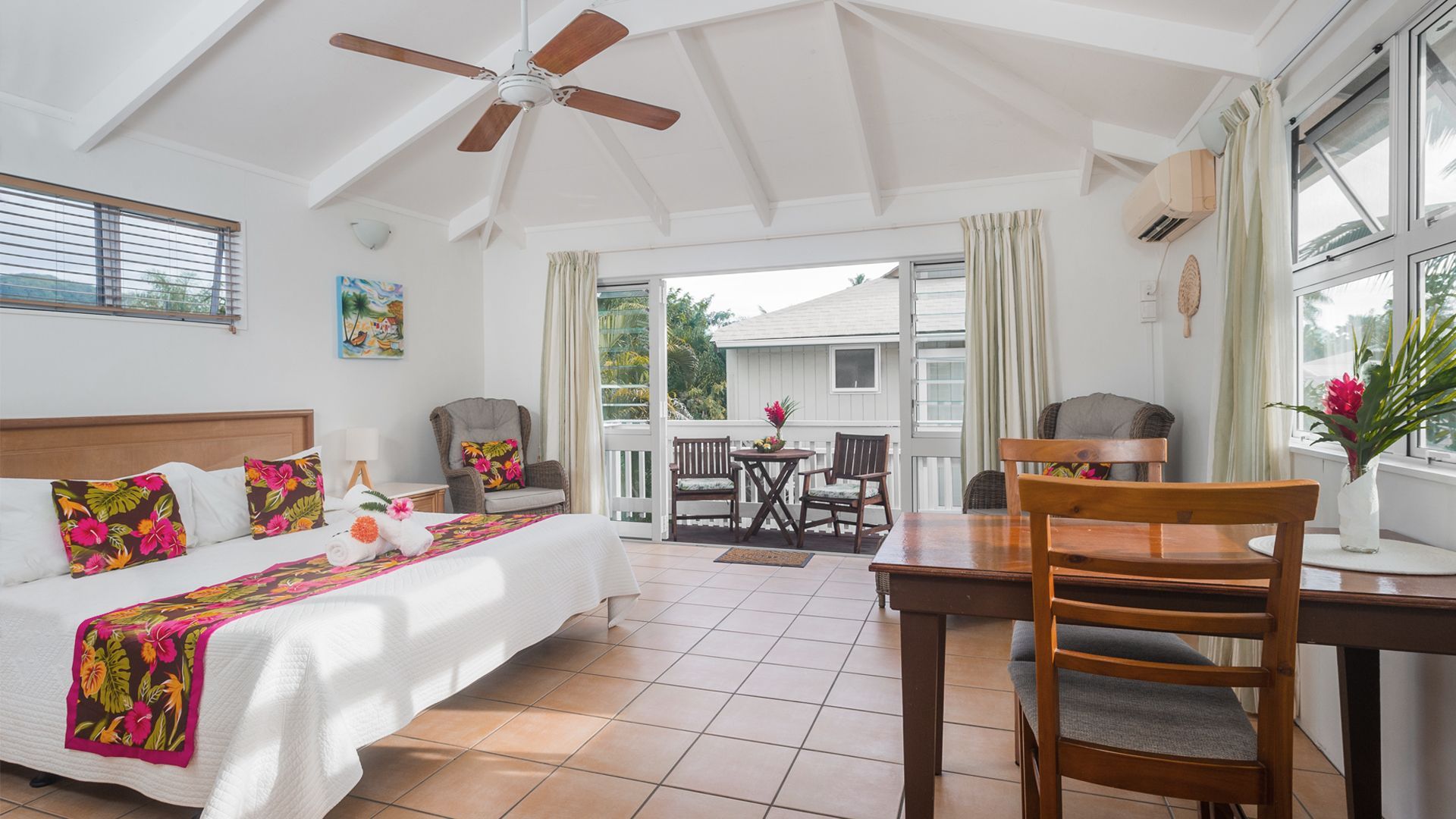 A bright hotel room with a bed featuring floral linens, tiled floors, a dining area, and a balcony view.