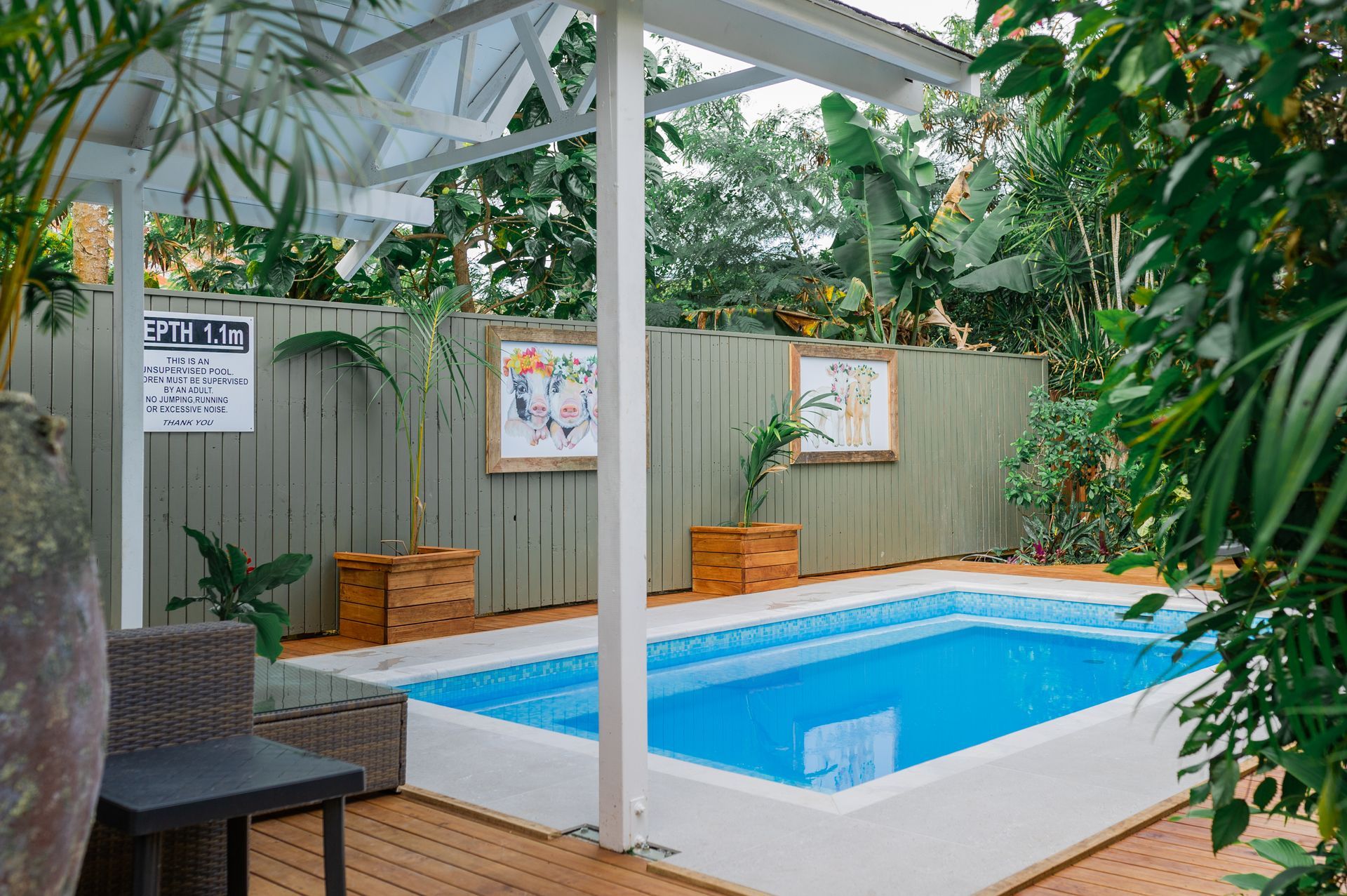 A small, rectangular blue swimming pool on a wooden deck, partially shaded by a white canopy and surrounded by greenery.