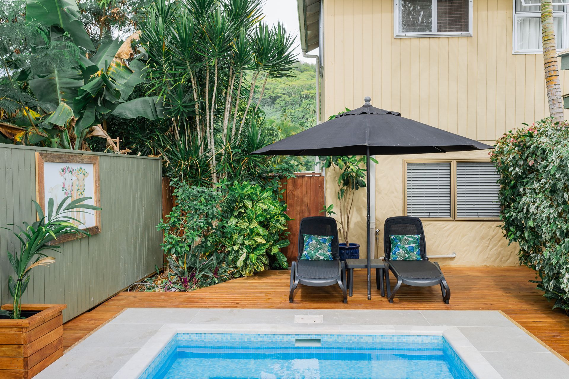 Two lounge chairs with cushions sit under a black umbrella on a wooden deck next to a small rectangular swimming pool.