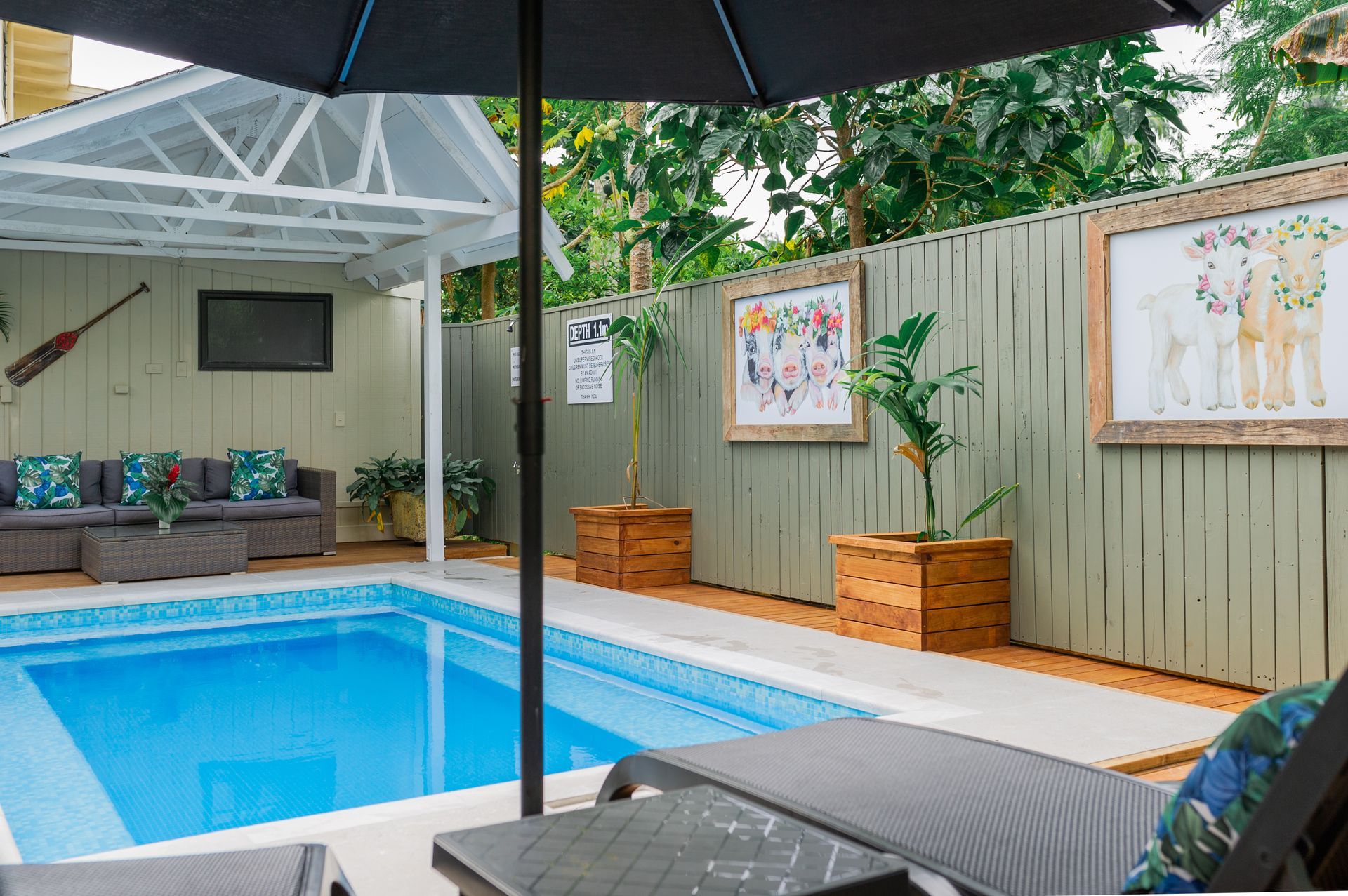 A backyard pool area featuring lounge seating, an umbrella, wooden planters, and decorative wall art on a pale fence.