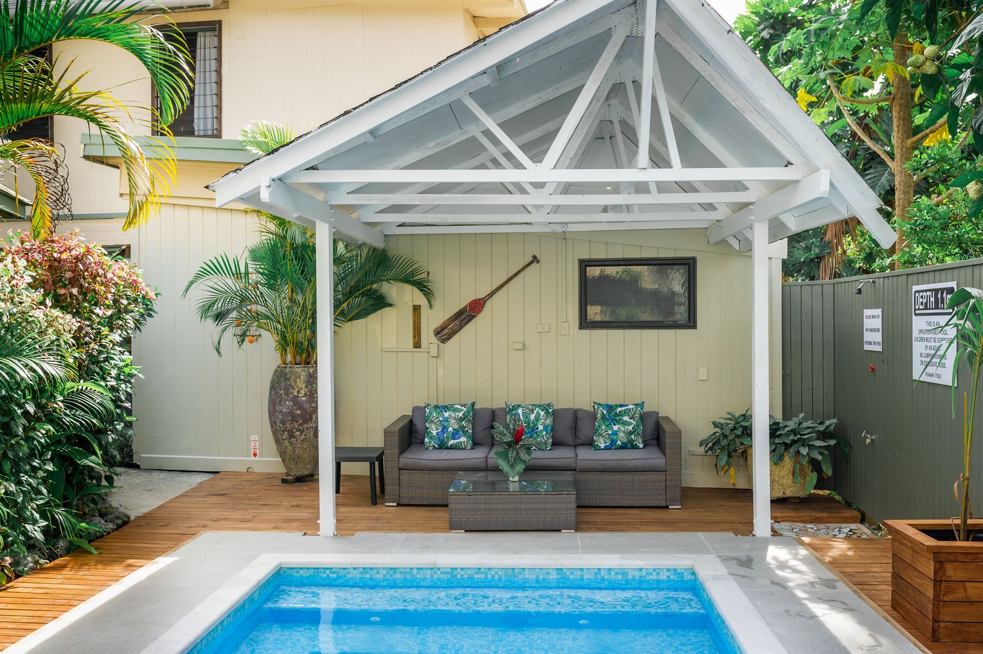 A poolside lounge area under a white pergola with a grey sofa, small coffee table, and a decorative oar on the wall.