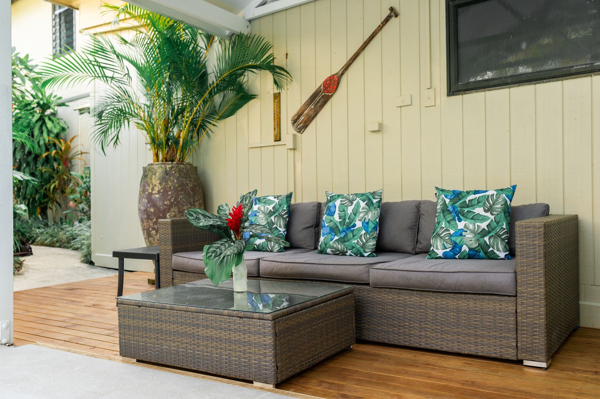 A patio lounge area features a grey wicker sofa with tropical-patterned pillows and a glass-topped table near a potted palm.