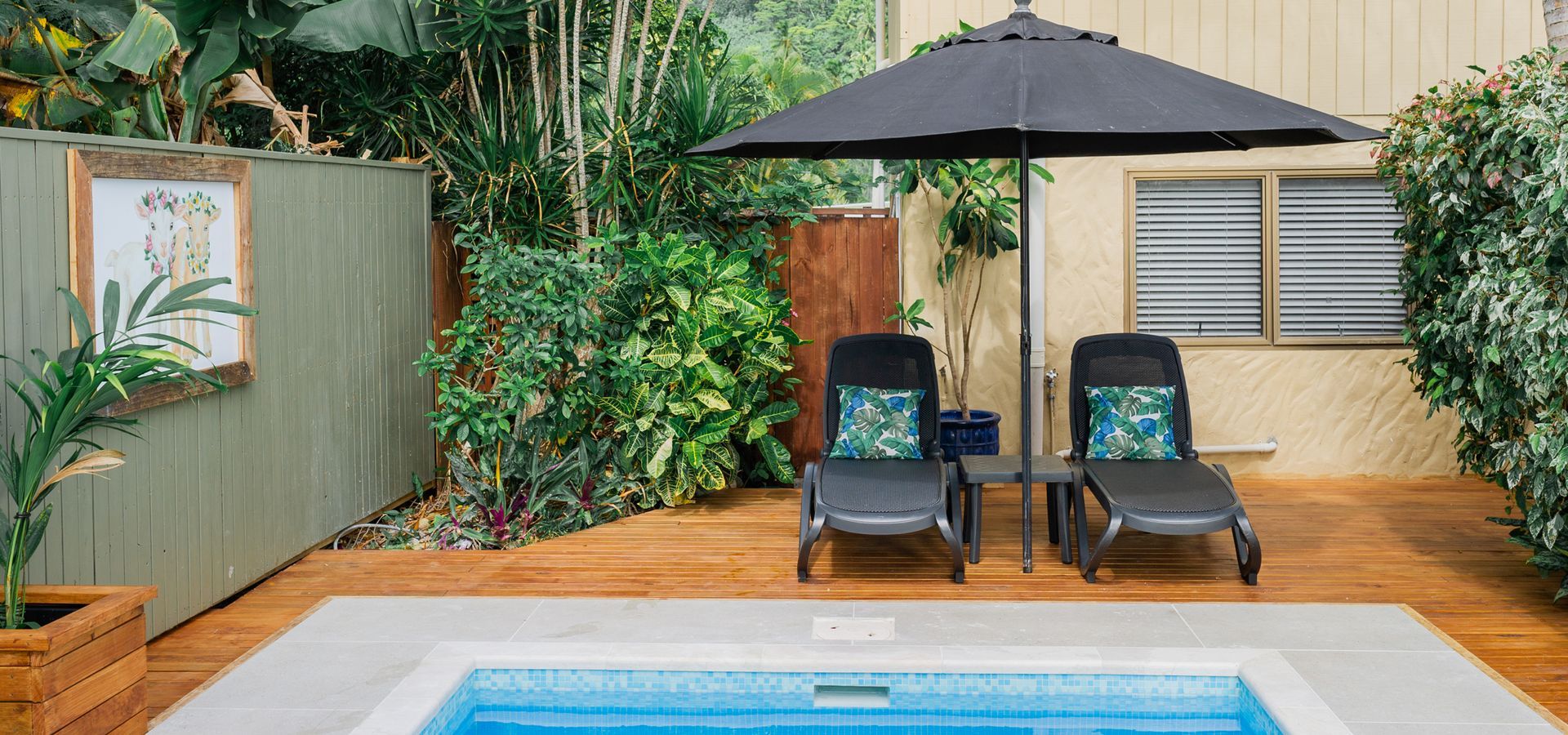 Two lounge chairs with teal cushions under a black umbrella on a wooden deck next to a backyard pool.
