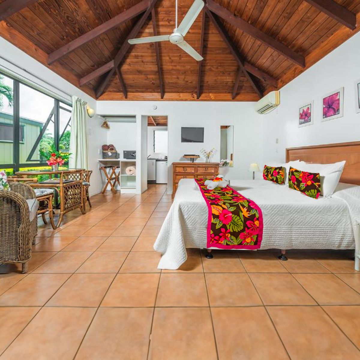 A hotel room with high wood-beamed ceilings, tile floors, a white bed with a floral runner, and a wicker chair set.