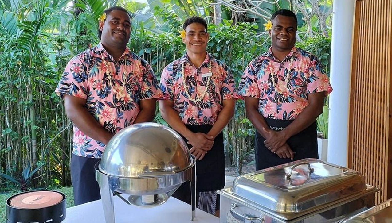 Three hotel staff in matching floral shirts stand behind a buffet table with stainless steel serving dishes outdoors.