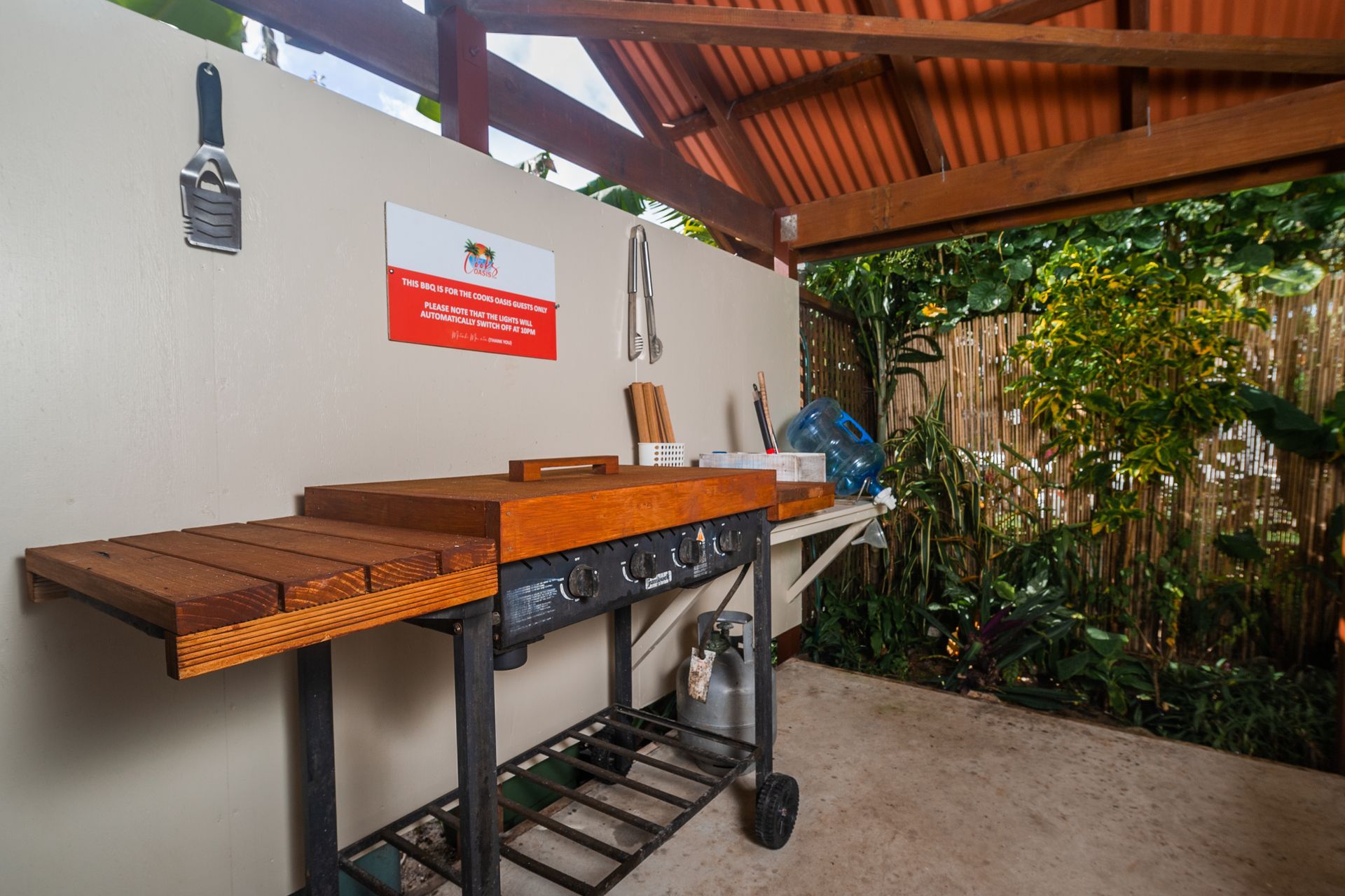 An outdoor grilling station with a wooden-topped grill, a gas canister, and wall-mounted tools under a covered patio.