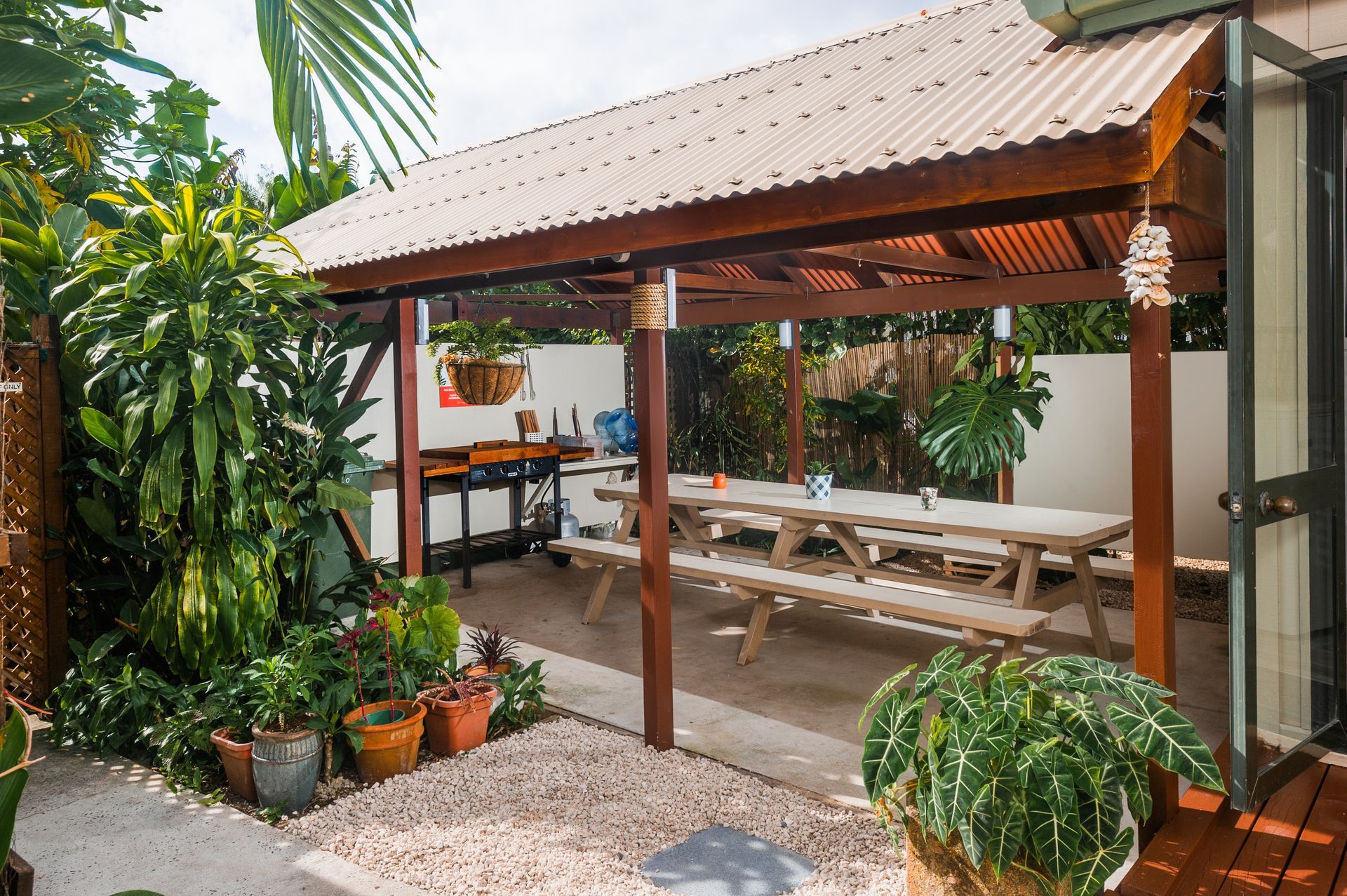 Outdoor dining area with a wooden gazebo, a long picnic table, and various potted plants on a gravel patio.