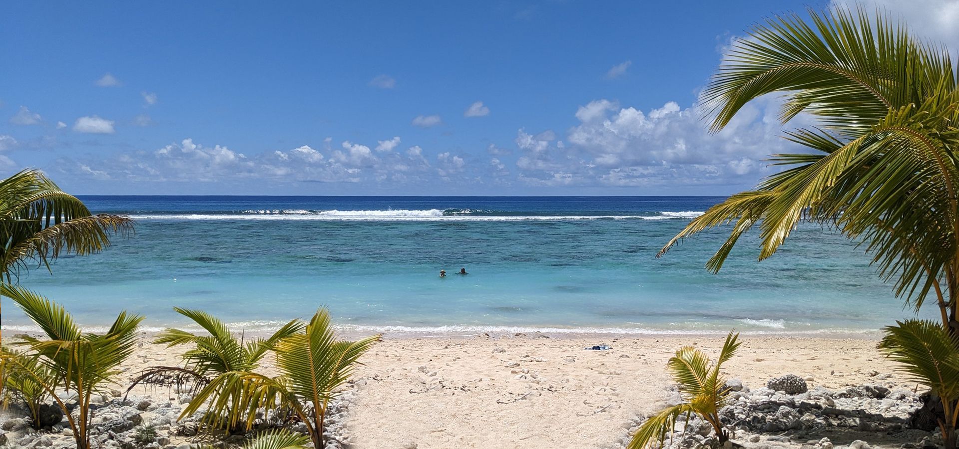 A tropical beach scene with white sand, palm trees, turquoise shallow water, and a dark blue ocean under a sunny sky.