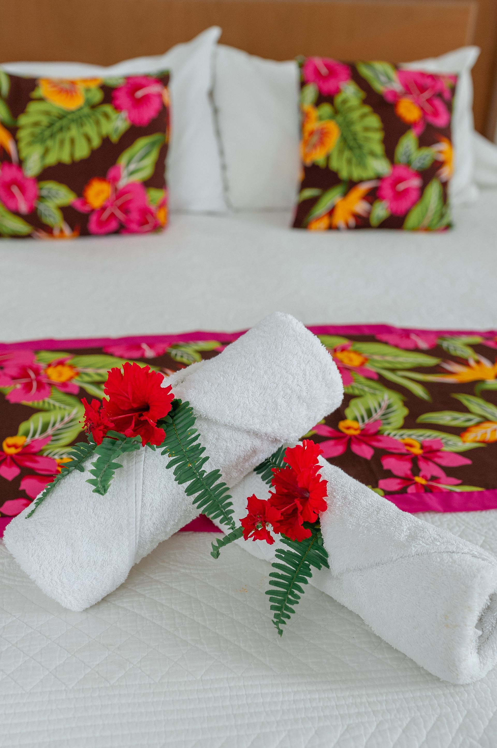 Two rolled white towels decorated with red flowers and green ferns on a bed with floral pillows and a matching runner.