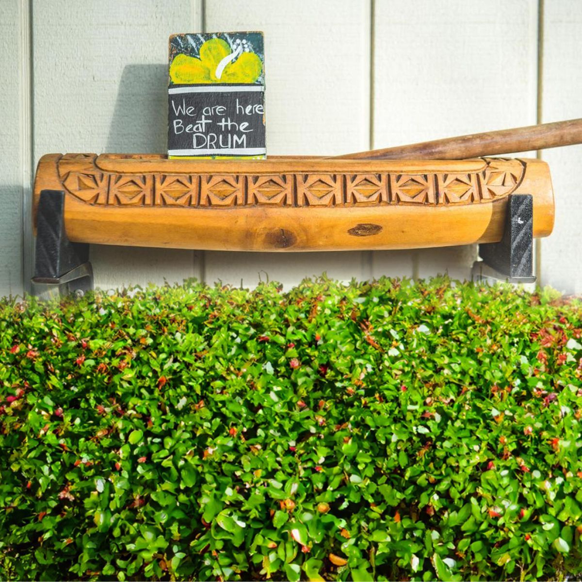 A carved wooden log drum rests on black supports behind a lush green bush, with a sign reading