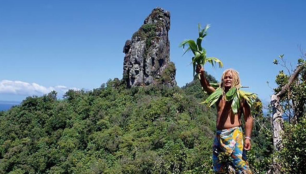 A person with blonde hair stands in a green, mountainous landscape holding a leafy plant against a prominent rock spire.
