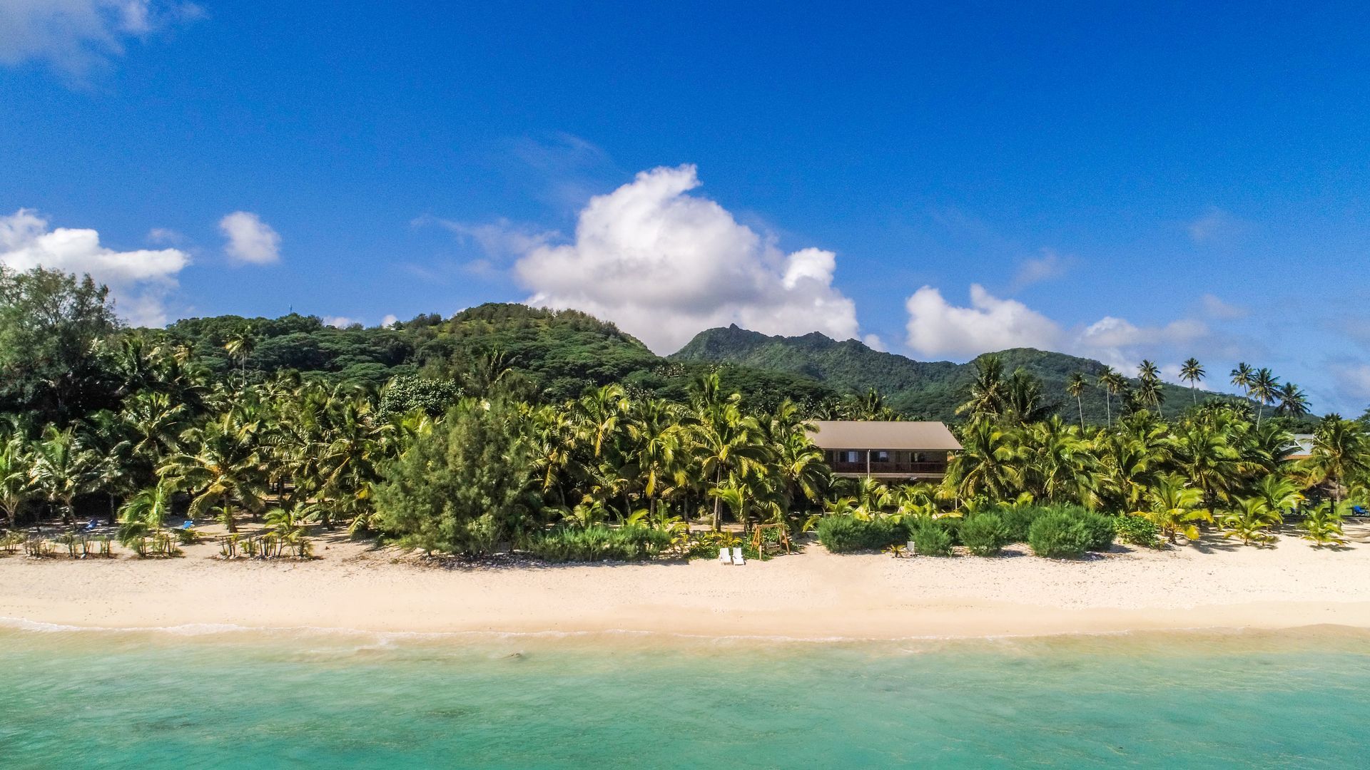 A sandy beach meets turquoise water, backed by lush green palm trees and rolling hills under a bright blue sky.
