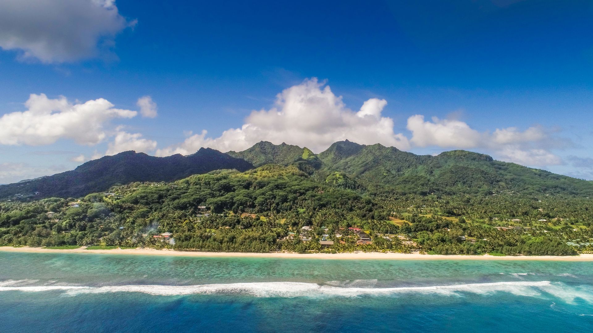 Lush green mountains rise above a sandy beach and turquoise reef under a bright blue, partly cloudy sky.