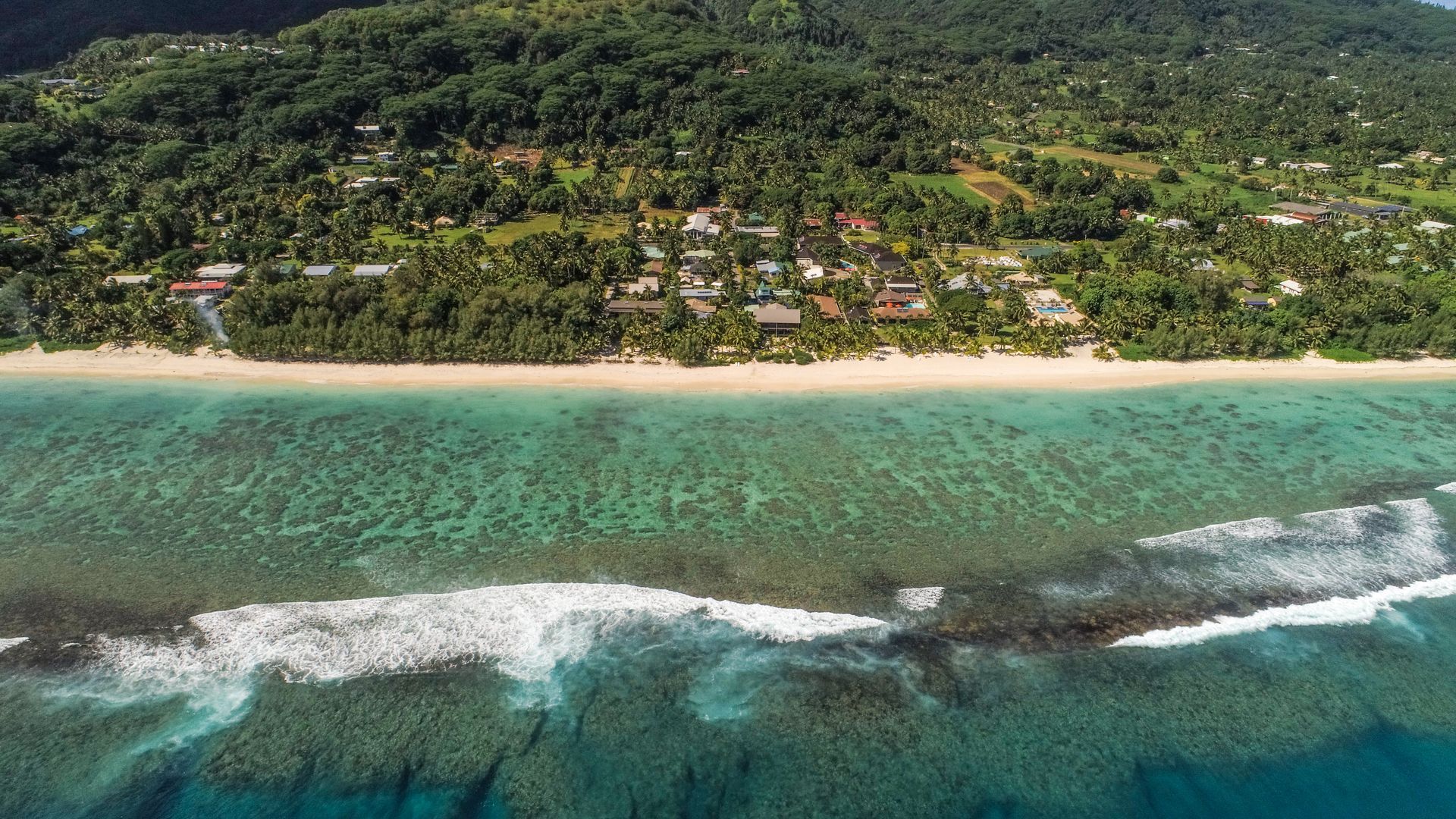 Aerial view of a white sand beach with turquoise reef waters, waves breaking on the reef, and a lush, tree-filled coast.