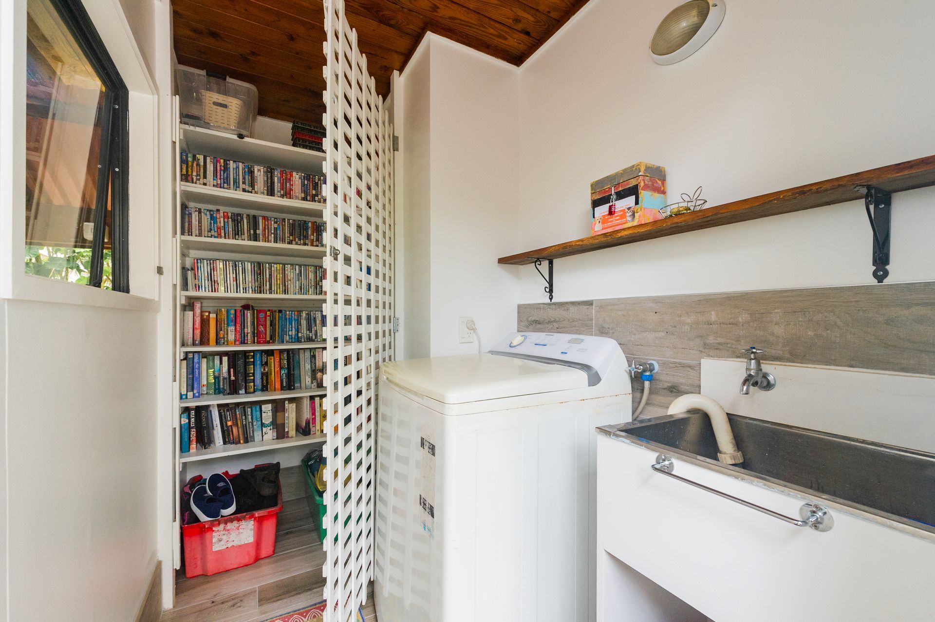 A utility room with a white washing machine, a metal sink, a wall shelf, and a shelving unit packed with many books.
