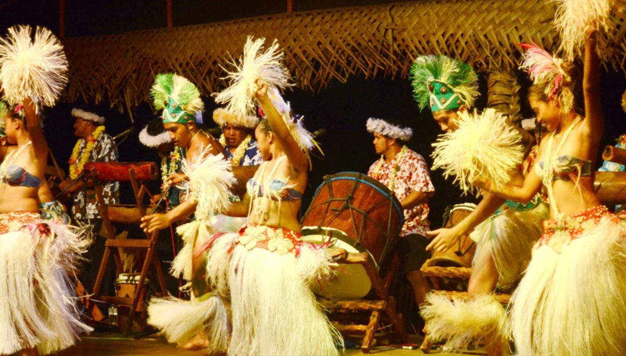 Performers in traditional Polynesian dance attire with grass skirts and headdresses dance on stage with musicians nearby.