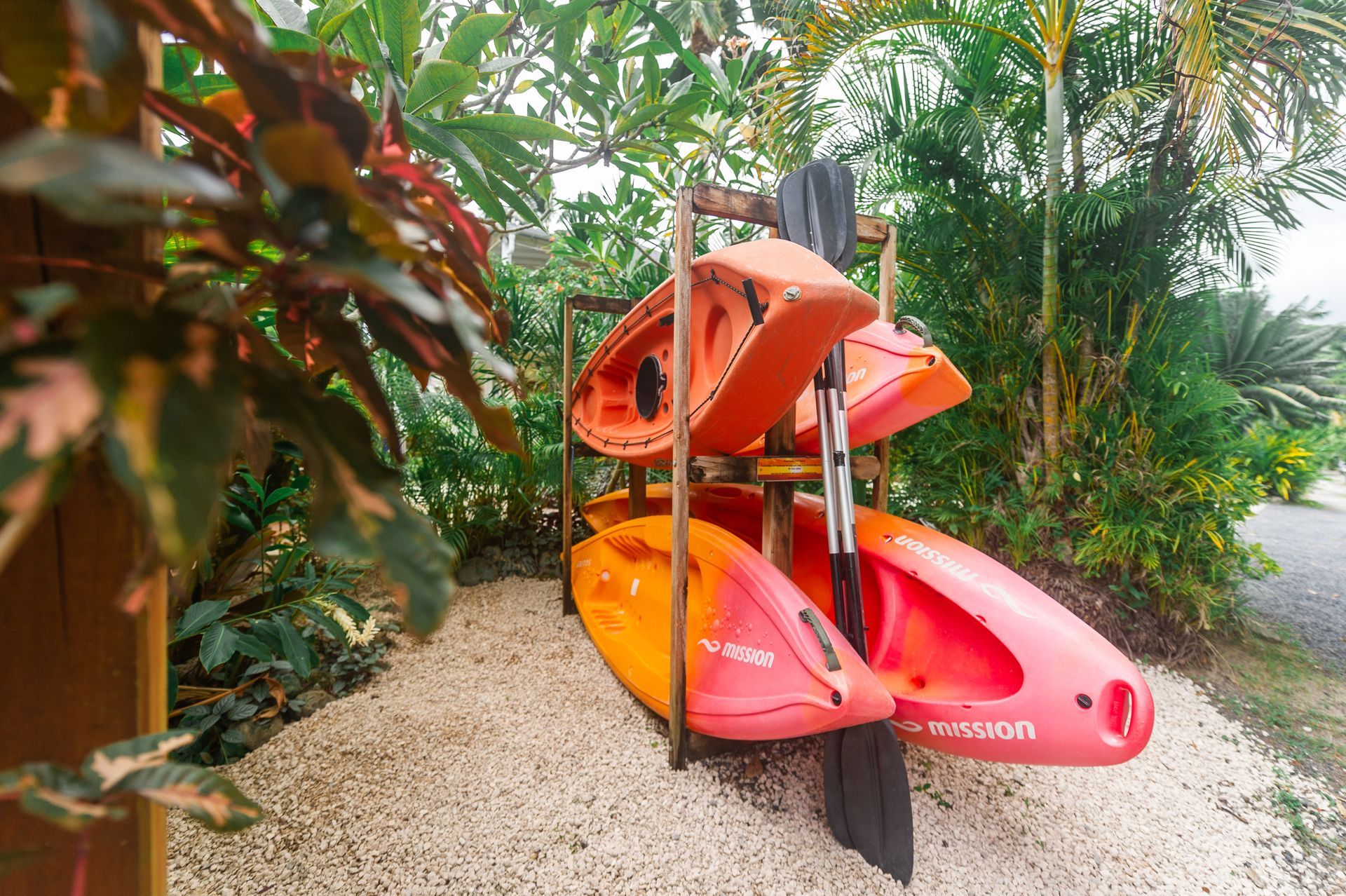 Two orange and yellow kayaks stored on a wooden rack outdoors, with a paddle leaning against them on a gravel path.