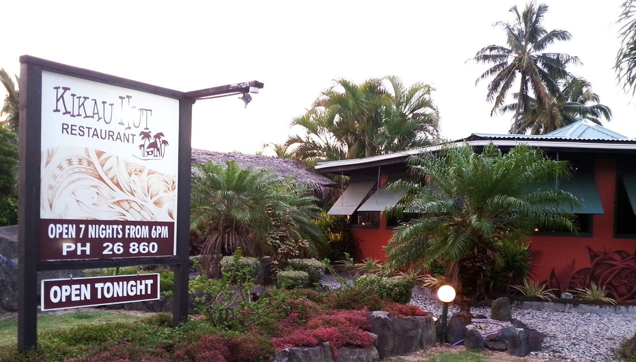 A sign for the Kirau Hut Restaurant stands in front of a red building surrounded by lush greenery and palm trees.