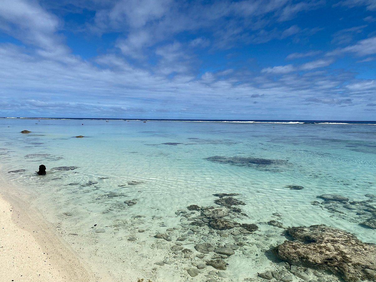 A clear, shallow tropical lagoon with rocky coral formations under a bright blue sky with light clouds.