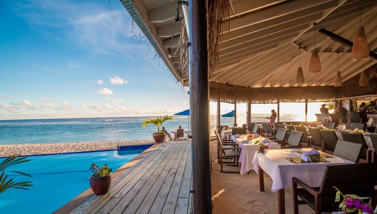A restaurant terrace overlooking a blue ocean with an infinity pool under a thatched roof at sunset.