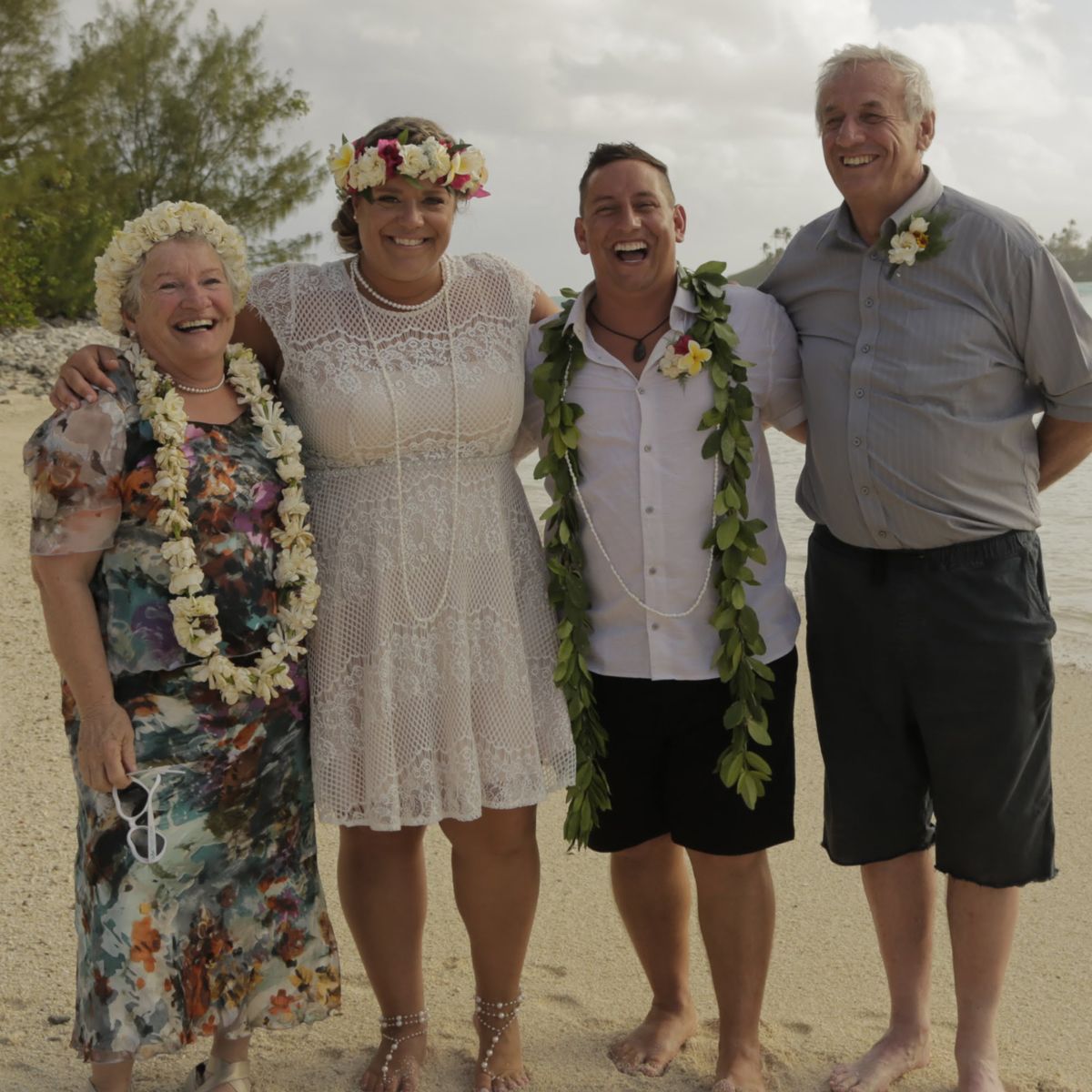 Four people stand on a sandy beach, smiling and wearing floral leis, at a casual outdoor wedding celebration.