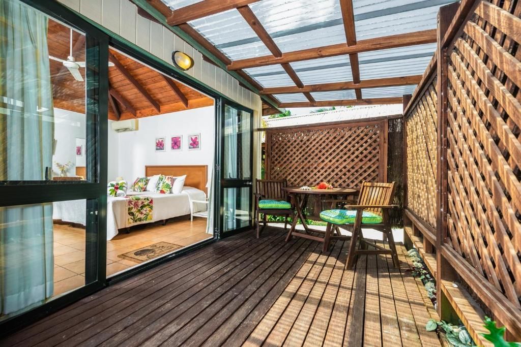 A wooden patio deck with a small table and chairs, covered by a slatted roof, leading into a bedroom with a double bed.