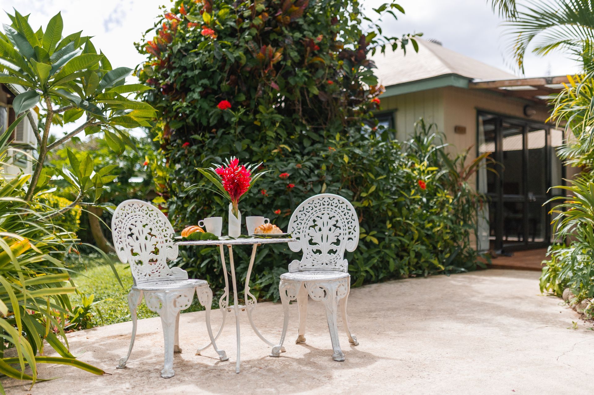 A patio set with two white metal chairs and a table featuring a vase of red flowers sits on a gravel path near a house.
