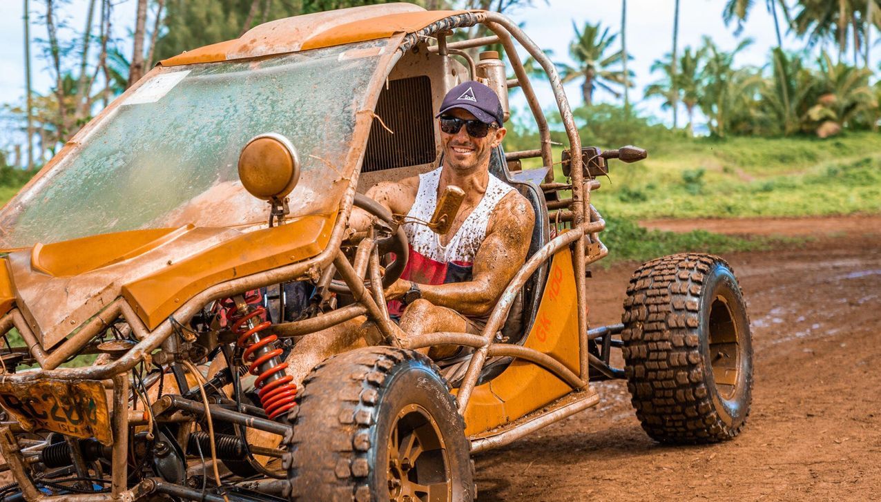 A person wearing sunglasses and a cap driving a mud-splattered off-road buggy on a dirt trail.