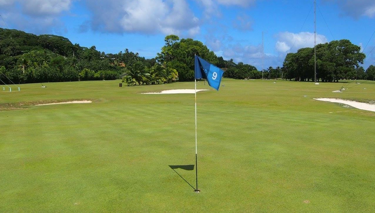 A blue golf flag marked with the number 9 stands on a green in the foreground, with sand traps and trees in the distance.