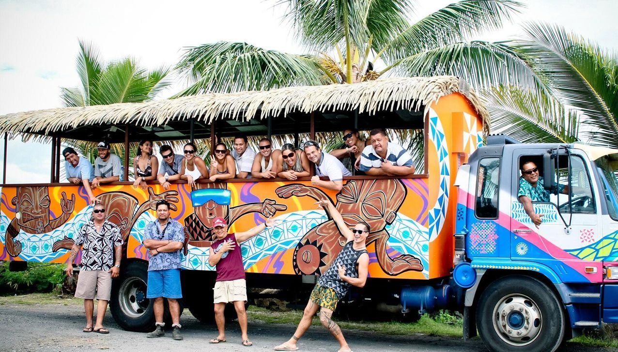 A group of people standing in front of and inside an orange, decorated open-air bus with a thatched roof.